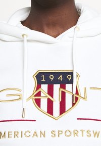 Felpa bianca con cappuccio, caratterizzata da un logo scudo ricamato in oro e rosso con il testo "GANT" e "1949". I laccetti hanno estremità in metallo dorato.