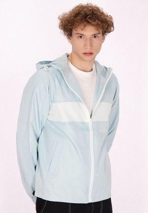 Giacca zip-up azzurro chiaro con una striscia orizzontale bianca, cappuccio regolabile e due tasche laterali. Texture liscia e design casual.