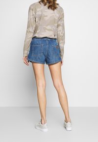 Pantaloncini in denim blu medio, con vita alta, orli sfrangiati e due tasche posteriori. Abbinati a una camicia mimetica a maniche lunghe.