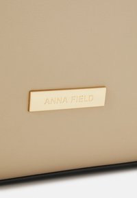 Beige Kunstledertasche mit glatter Oberfläche, ausgestattet mit einer goldenen rechteckigen Logo-Plakette, auf der "ANNA FIELD" deutlich eingraviert ist.