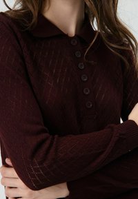 Bordeauxrode gebreide trui met een ruitpatroon, knoopsluiting bij de kraag en geribbelde manchetten. Voorzien van zes donkere knopen aan de voorkant.