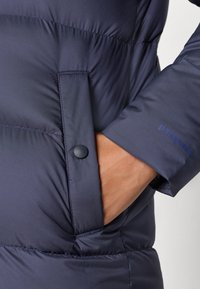 Tamsiai mėlynas pūkinis džemperis su lygiu, blizgiu paviršiumi, turintis kišenę su saga ir logotipą ant rankovės. Medžiaga atrodo lengva ir izoliuota.