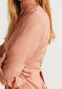 Kobieta z długimi blond włosami ubrana w brzoskwiniową bluzkę z długim rękawem, ze podwiniętymi rękawami i detalami w postaci guzików, z założonymi na siebie rękami.