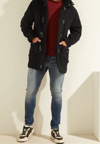 Parka negra con cierres de tipo toggle, dos bolsillos frontales y capucha, llevada sobre una camisa burdeos y jeans azules desgastados, combinada con zapatillas.