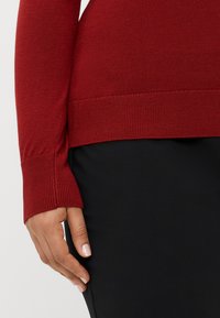 Ilgi piedurkņu sarkans džemperis izsmalcinātā adījumā, ar ribotām aprocēm un apakšmalu. Saskaņots ar gludu melnu svārku, uzsverot pieguļošo siluetu.