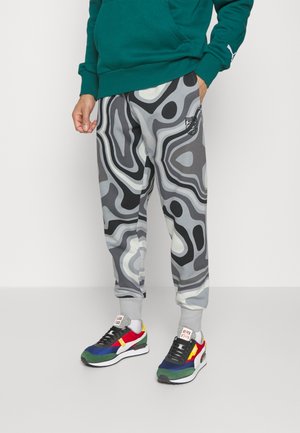 Personne portant un pantalon de jogging Puma gris et noir à motif abstrait, un sweat-shirt vert et des baskets multicolores avec des chaussettes blanches.