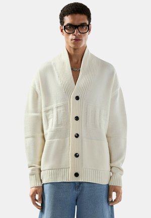 Uomo che indossa un cardigan oversize color crema con bottoni neri e lettere in rilievo, abbinato a jeans azzurri e occhiali neri.