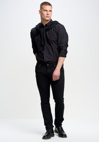 Camisa negra de botones con mangas largas, jeans negros y zapatos negros con cordones. Un suéter negro está drapeado sobre los hombros.