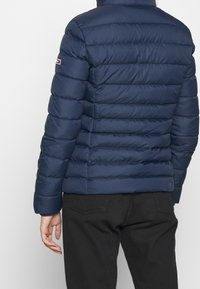 Back view of a person wearing a navy blue puffer jacket with horizontal quilting and black pants standing against a plain background.