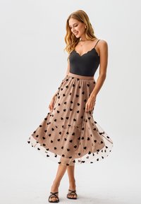 Schwarzer figurbetonter Body mit einem wellenförmigen Ausschnitt, kombiniert mit einem durchsichtigen braunen Rock mit schwarzen Herzmustern. Schwarze Riemchensandalen.