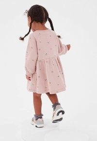 Kind in einem rosa Kleid mit Tiermustern, das auf einem weißen Podest steht, bunte Sneaker trägt und zwei Zöpfe im Haar hat.