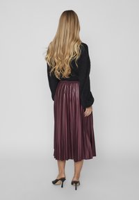 Suéter negro de punto, falda midi plisada morada, textura fluida, combinada con tacones negros. El modelo se ve desde atrás sobre un fondo claro.