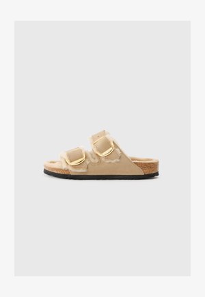 Tan slide sandals with two adjustable buckle straps, soft faux fur accents, and a cork footbed, featuring a textured black rubber sole.