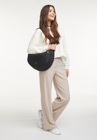 Borsa a tracolla nera in nylon con forma curvata, caratterizzata da un logo discreto e una singola tracolla regolabile, abbinata a pantaloni beige morbidi e sneakers bianche.