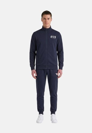 Giovane uomo che indossa una giacca blu navy con zip e pantaloni jogger abbinati con sneaker bianche, in piedi davanti a uno sfondo semplice.