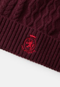 Bordeaux gebreide beanie met een kabelpatroon, een dikke geribbelde manchette en een rood geborduurd logo met een kroon en leeuw ontwerp.