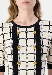 Bluză cu un model pe un fond crem, cu un grid negru și accente volănele negre. Dispune de nasturi aurii pe toată lungimea frontală și un decolteu rotund.