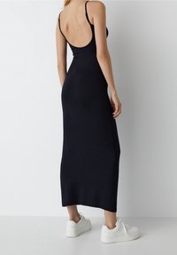Robe midi noire ajustée avec des fines bretelles, design à dos nu, texture lisse, associée à des baskets blanches. Silhouette simple et épurée.