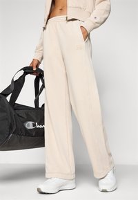 Beige sweatpants med en avslappnad passform och elastisk midja, som har sidofickor och diskret logobroderi, i kombination med en svart duffelväska.