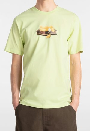 Homme portant un t-shirt vert clair avec un motif d'horloge digitale affichant "19:66" et un petit animal en peluche jaune posé dessus.