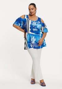 Tunika mit blauem Blumenmuster und kühlen Schultern, kombiniert mit weißen Schlaghosen und blauen Riemchensandalen. Glattes Material, lässige Passform.