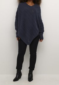 Mörkblå stickad poncho med V-ringning, med diagonala ribbade mönster och öppna vävdetaljer. Bärs över svarta åtsittande byxor och stövlar.