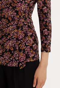 Blommig omslagstopp i svart med rosa och guldaccents. Har långa ärmar, en knytning i midjan och en subtil glans i tyget.