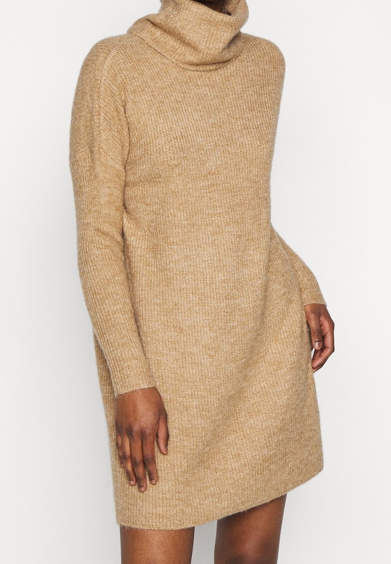 Personne portant une robe-pull à col roulé en tricot beige à manches longues avec un motif texturé, debout sur un fond blanc uni.