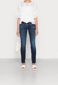 Blusa bianca e trasparente con ricami floreali e maniche arricciate, abbinata a jeans skinny blu scuro e sandali bianchi con cinturini; tiene una borsa all'uncinetto.