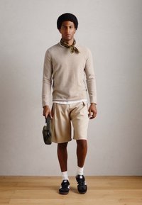 Beiger Pullover, braune Shorts, schwarze Turnschuhe, weiße Socken und ein grüner Schal. Hält eine grüne Tasche. Einfaches Design, legere Bekleidung.