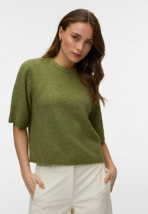 Femme aux longs cheveux bruns portant un pull vert à manches courtes et un pantalon blanc, les mains dans les poches, debout devant un fond uni.