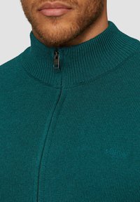 Türkiser Pullover mit Reißverschluss und geripptem Textur, ausgestattet mit einem schwarzen Reißverschluss und einem dezenten Logo am Saum. Weicher Strickstoff.