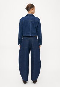 Moteris su susuktais plaukais, vilkinti tamsiai mėlyną džinsinį trumpą švarką ir platėjančiomis kelnėmis, stovi su nugara nusigręžusi į žiūrovą baltoje fone.