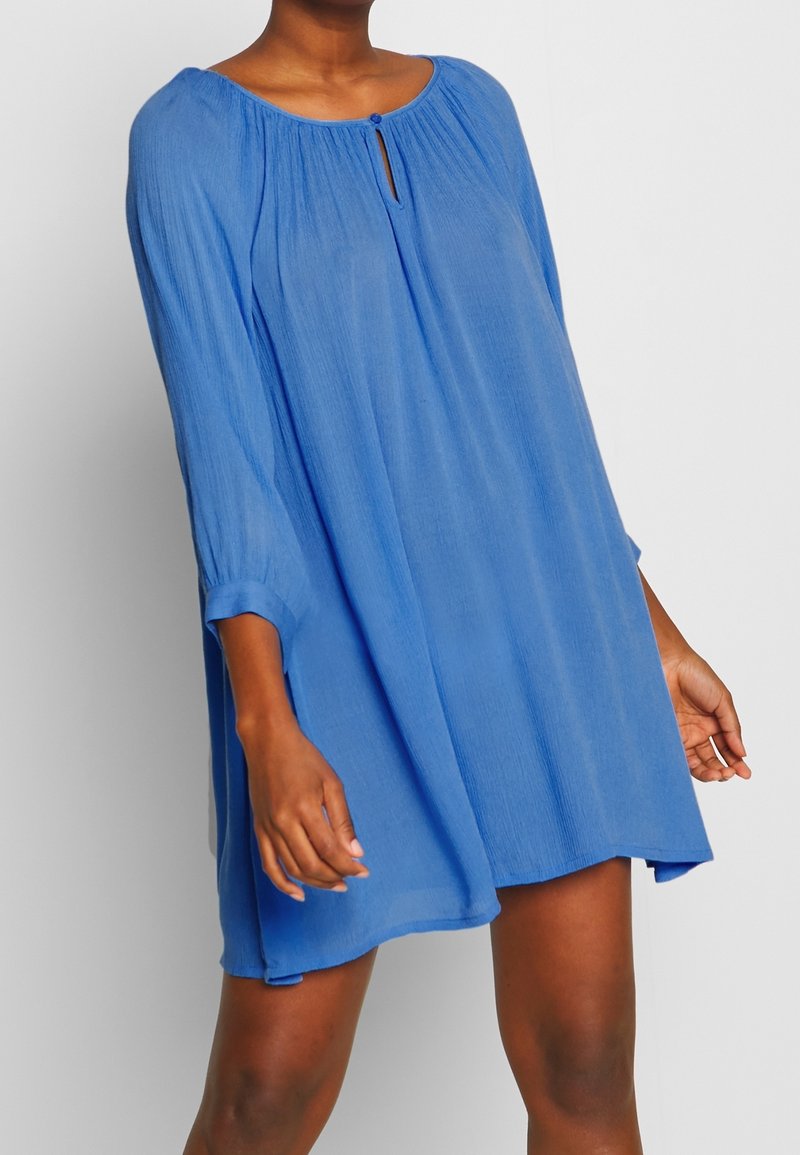 Femme portant une robe bleue ample à manches longues avec une petite ouverture en forme de goutte avec une fermeture à bouton, debout devant un fond uni.