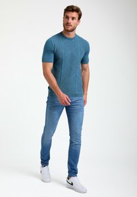 Mėlynas trumpomis rankovėmis marškinėliai su geometrine tekstūra, derinami su šviesiai mėlynomis džinsais ir baltais sportbačiais, turinčiais juodą swoosh emblema.