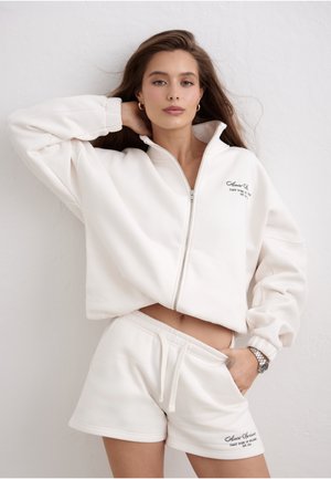 Kvinde med langt brunt hår iført hvid lynlås-sweatshirt og matchende shorts, stående mod en hvid tekstureret væg.