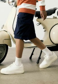 Shorts cargo bleu marine, chaussettes blanches à la cheville et baskets blanches portés par une personne marchant à côté d'un scooter vintage crème sur un sol en béton.