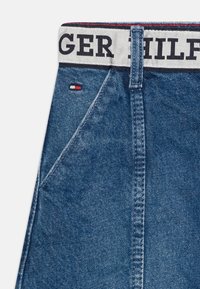 Pantalones cortos vaqueros con una cinturilla blanca que lleva el texto "TOMMY HILFIGER", tela azul con textura sutil y una pequeña etiqueta de logotipo en rojo y blanco.