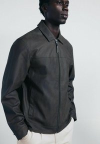 Giacca di pelle nera con zip frontale, colletto a punta e tasche laterali, dal texture liscia e design minimalista.