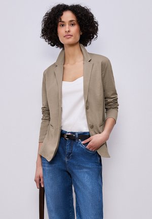 Mujer con cabello rizado, vestida con blazer beige, camiseta blanca, jeans azules y cinturón negro, de pie con una mano en el bolsillo y sujetando la correa de una bolsa.