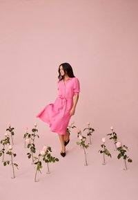 Femme en robe rose marchant parmi des vases en verre tenant des roses roses sur un fond rose, souriante et regardant de côté.