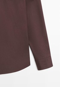 Poignet de chemise à manches longues marron foncé avec un simple détail de couture et un ourlet arrondi sur fond blanc.