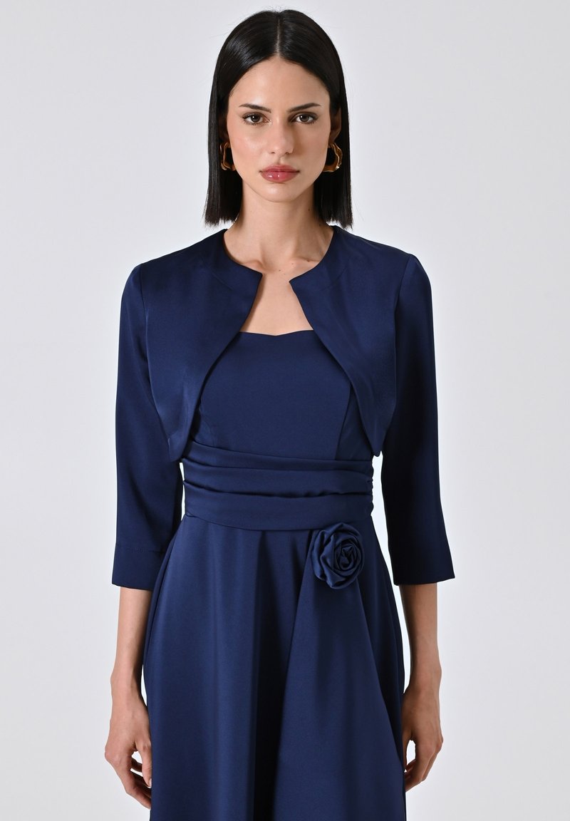 Abito blu navy con corpino aderente e dettaglio a forma di rosa in vita, abbinato a un giacchetto corta strutturato nello stesso tessuto.