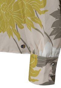 Seidenbluse mit großen gelben Blumenmustern auf einem hellbeigen Hintergrund, mit einer Manschette und Knopfdetails in einem kontrastierenden Farbton.
