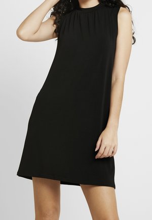 Kobieta ubrana w czarną, bezrękawną, sięgającą kolan sukienkę z wysokim dekoltem, pozująca z jedną ręką przy głowie, a drugą opuszczoną wzdłuż ciała.