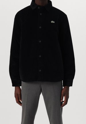 Mann trägt eine schwarze, knöpfbare Cordjacke mit einem grünen Krokodil-Logo auf der Brust und graue Hose.