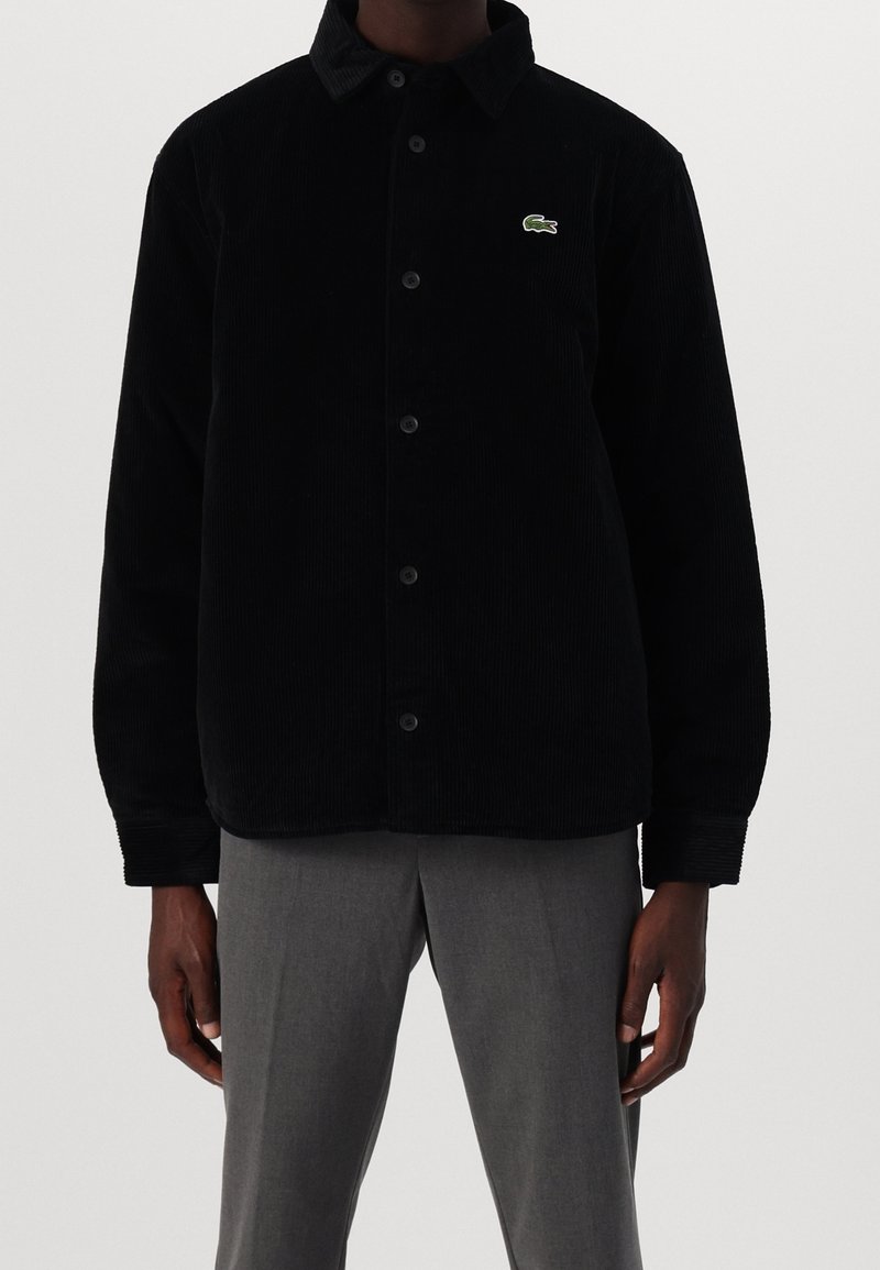Homme portant une veste en velours côtelé noire boutonnée avec un logo crocodile vert sur la poitrine et un pantalon gris.