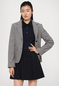 Harmaa ja musta ruudullinen bleiseri, jossa on notched-lapels, yhdistettynä tumman väriseen polo-paitaan ja mustaan pleated-hameeseen, jossa on pystysuorat viivat.