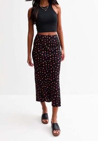 Kvinde der går iført en sort ærmeløs crop top, sort midi nederdel med små røde blomster, og sorte slid-sandaler.