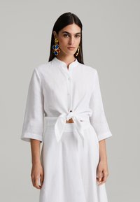 Bela lanena srajca z mandarin ovratnikom, zavezana v pasu. Dolgi rokavi z zavihanimi manšeti. Model nosi barvite, velike cvetlične uhane.